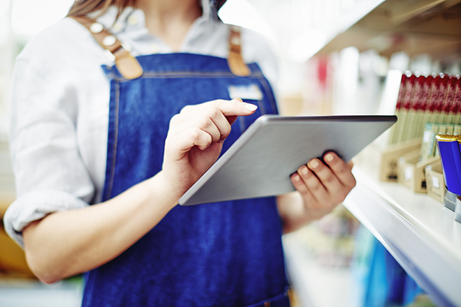 Midsection of deli owner touching digital tablet by shelves in store. Young saleswoman is working on wireless technology at shop. She is wearing denim overalls.