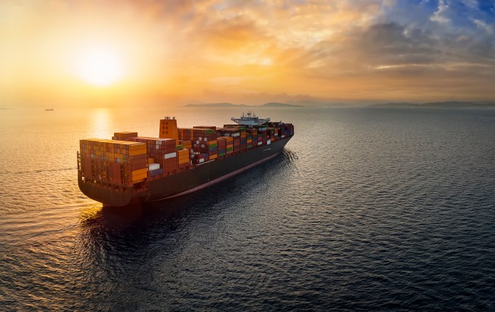 A large container ship sailing across the sea with the sun setting behind it.