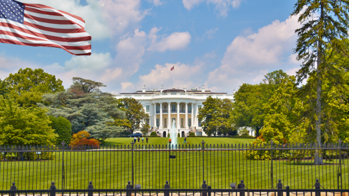 Image of the Whitehouse in the United States with the green lawn in front and the fountain elegantly displaying a jet of water. To the left hand side a Stars and Stripes flag is blowing in the breeze.