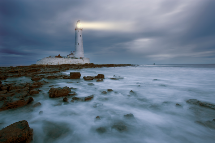 Image of a white stone lighthouse on a rocky seashore with sea mist on the waves. The sky is dark, potentially before daybreak and the light of the lighthouse shines out to sea.